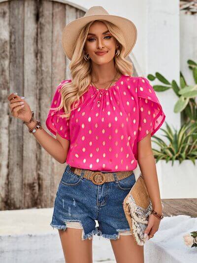 Model wearing the Amy Petal Sleeve Top in pink with gold accents, styled with denim shorts and a hat.