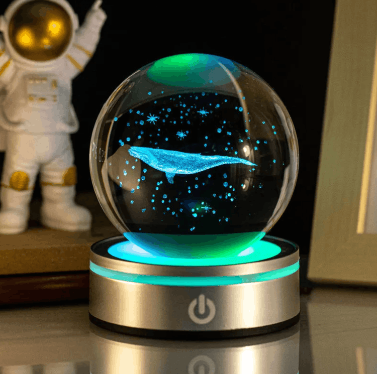 Glass ball with a whale inside, illuminated by green light, on a reflective surface.
